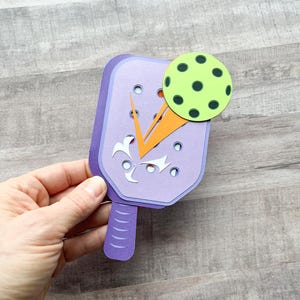 May include: Purple paper craft paddle with an orange and white design and a green and black paper ball.
