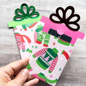 May include: Two pink gift card holders with a green and pink sweater pattern. The holders have a green and a pink bow on top. A gift card is visible in the pink holder.