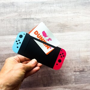 Peut inclure: Un porte-cartes noir con&ccedil;u pour ressembler &agrave; une console de jeu Nintendo Switch, avec des accents bleus et rouges. Une carte-cadeau Dunkin' est ins&eacute;r&eacute;e dans le porte-cartes. Le porte-cartes est tenu dans une main.