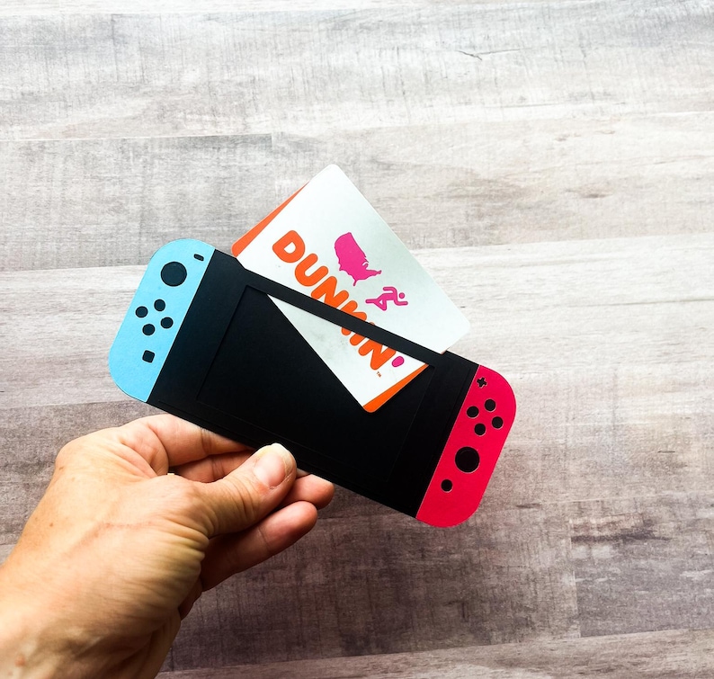 Peut inclure: Un porte-cartes noir con&ccedil;u pour ressembler &agrave; une console de jeu Nintendo Switch, avec des accents bleus et rouges. Une carte-cadeau Dunkin' est ins&eacute;r&eacute;e dans le porte-cartes. Le porte-cartes est tenu dans une main.