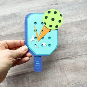 May include: A blue and orange paper craft of a pickleball paddle and ball. The paddle has a blue handle and a light blue face with orange and white details. The ball is green with black dots.