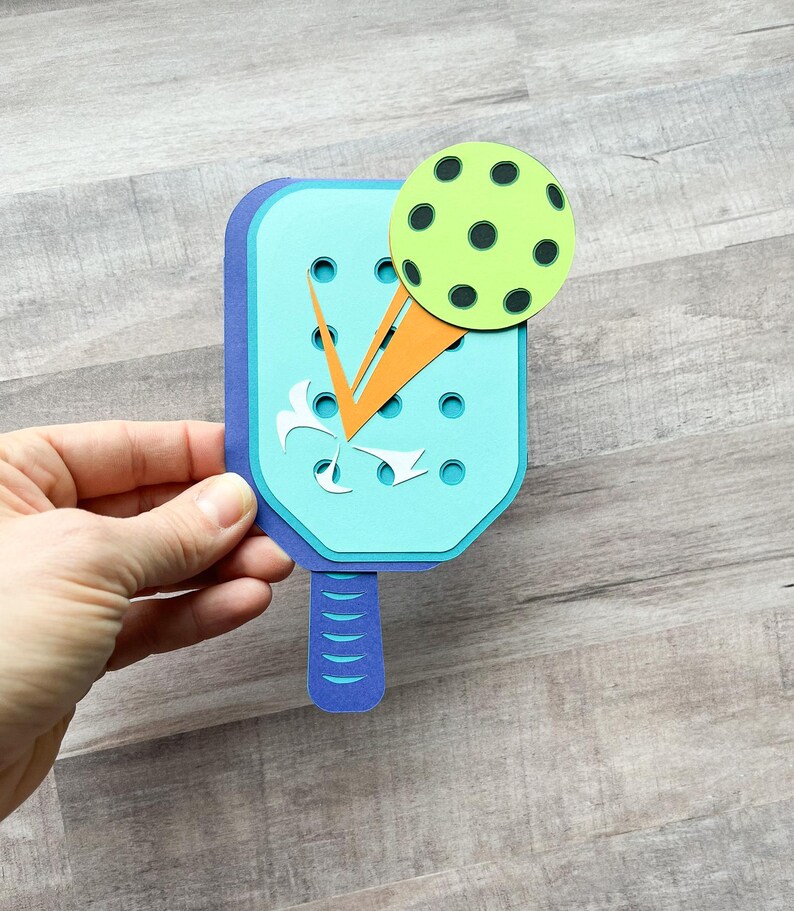 May include: A blue and orange paper craft of a pickleball paddle and ball. The paddle has a blue handle and a light blue face with orange and white details. The ball is green with black dots.
