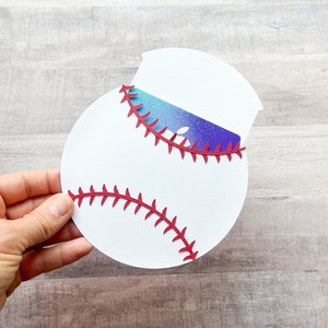 May include: A white circular cardstock cutout shaped like a baseball with red die-cut stitching details. A rectangular gift card with a blue and purple galaxy-themed gradient and a white apple logo is inserted into a slot at the top of the card.