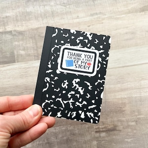May include: A small black and white composition notebook with a sticker that reads "THANK YOU FOR BEING A PART OF MY STORY". The sticker features a blue book and a red apple.