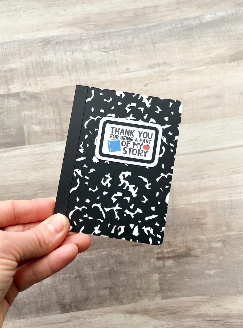 May include: A small black and white composition notebook with a sticker that reads "THANK YOU FOR BEING A PART OF MY STORY". The sticker features a blue book and a red apple.