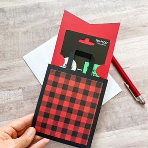 May include: A gift card holder with a black and red buffalo plaid pattern. The holder has a black border and a small handle. A red card and a gift card are visible inside. A red pen and a white envelope are also in the image.