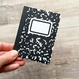 May include: A small, black and white composition notebook with a classic design. The cover features a black background with white abstract shapes and a white label area. The notebook is held in a hand.