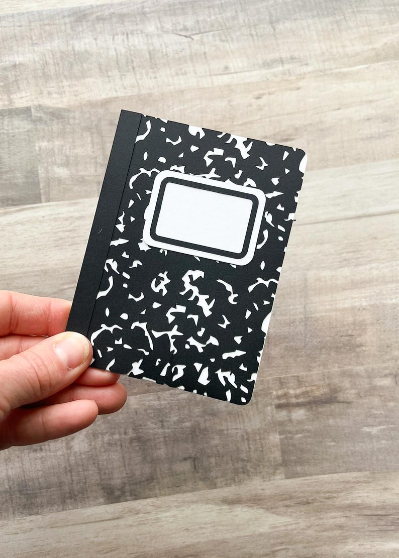 May include: A small, black and white composition notebook with a classic design. The cover features a black background with white abstract shapes and a white label area. The notebook is held in a hand.