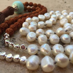 May include: A brown tassel hangs from a mala necklace made with brown beads, white pearls, and silver beads. The necklace has a turquoise bead at the top.