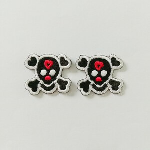 Könnte beinhalten: Zwei aufgestickte Aufnäher mit einem Totenkopf-Design. Die Totenköpfe sind schwarz mit weißen Akzenten, roten Herzen und roten Mündern. Die Aufnäher befinden sich auf weißem Hintergrund.