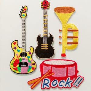 Könnte beinhalten: Gestickte Aufnäher mit Musikinstrumenten: eine gelb-schwarze Gitarre mit bunten Akzenten, eine schwarze Gitarre, eine gelbe Trompete und eine rote Trommel mit Trommelstöcken. Das Wort "Rock!!" ist darunter gestickt.