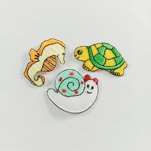 May include: Three embroidered fabric patches featuring a yellow and orange seahorse, a yellow and green turtle with a patterned shell, and a white snail with a light blue spiral shell decorated with small red dots and a red bow on its head.