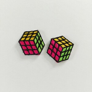 Puede incluir: Dos parches bordados con forma de cubos de rompecabezas. Cada parche presenta un patrón de cuadrícula con cuadrados de color amarillo brillante, rosa intenso y verde lima, delineados con hilo negro. El bordado tiene un aspecto texturizado.