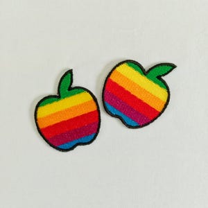 May include: Two embroidered apple patches with a rainbow striped design. Each apple has a green stem and a black outline. The rainbow stripes include red, orange, yellow, green, blue, and purple.
