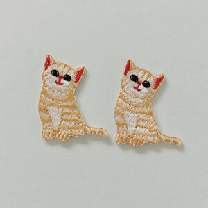 May include: Two embroidered patches featuring a seated cartoon cat. The cats have orange and white striped bodies, black eyes, red ears, and red smiles. The patches are likely for clothing or accessories.
