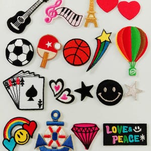 Puede incluir: Una colección de parches bordados con diversos diseños que incluyen una guitarra negra, teclas de piano, una torre Eiffel dorada, corazones rojos, un balón de fútbol, una paleta de ping pong, un balón de baloncesto, una estrella fugaz, un globo aerostático, cartas de juego, una cara sonriente, un ancla, un diamante y un parche rectangular con el texto LOVE & PEACE.