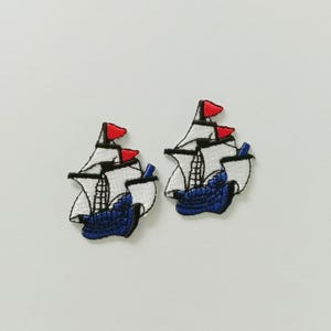 Puede incluir: Dos parches bordados de un velero con velas blancas, casco azul y banderas rojas. Los parches están detallados con costuras negras y están sobre un fondo blanco. Ideales para decorar ropa o accesorios.
