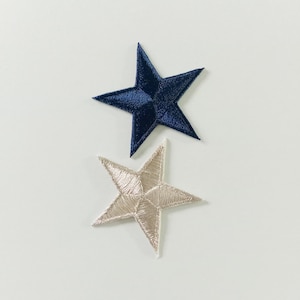 Puede incluir: Dos parches bordados con forma de estrella. Uno es azul marino, el otro es de color beige claro. Las estrellas tienen un diseño de cinco puntas y están hechas de tela texturizada. Probablemente sean para fines decorativos.