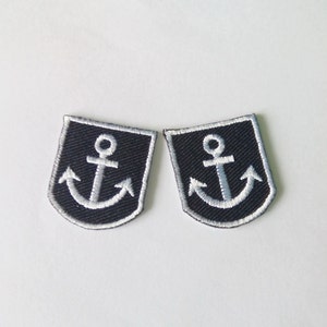 May include: Two navy blue anchor patches with white embroidered details. Each patch features a shield shape with a white anchor design. The patches are placed side by side on a white background.