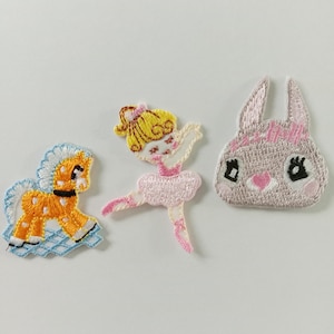 Puede incluir: Tres parches bordados: un caballo amarillo y naranja, una bailarina en rosa y un conejito gris con un lazo rosa. El caballo tiene una melena azul y está sobre una rejilla azul. La bailarina tiene el pelo amarillo y un tutú rosa. El conejito tiene ojos negros.