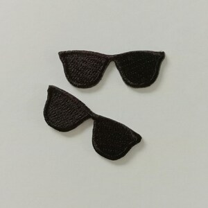 Puede incluir: Dos parches bordados negros con forma de gafas de sol. Los parches son de color marrón oscuro y tienen un aspecto texturizado. Las gafas de sol son de estilo clásico, con forma rectangular. Los parches están colocados sobre un fondo blanco.