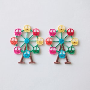 Può includere: Due toppe ricamate a forma di ruota panoramica. Ogni toppa presenta un design colorato con un centro turchese, una base marrone e una cornice verde. Le cabine della ruota panoramica sono rosa, gialle, rosse, arancioni e verdi.