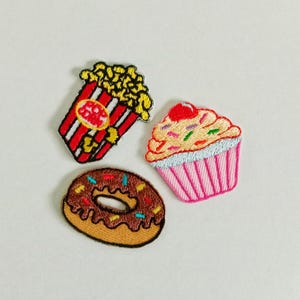 May include: Embroidered patches of a popcorn container with red and white stripes, a cupcake with pink frosting and a cherry, and a chocolate donut with sprinkles. Ideal for customising clothing or accessories.