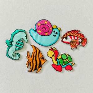 May include: A collection of five embroidered patches featuring aquatic creatures. Includes a teal seahorse, a light blue snail with a pink shell, a red and white fish, an orange fish with black stripes, and a turtle with a green shell and red feet.