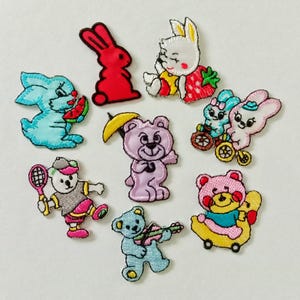 May include: A collection of embroidered patches featuring cartoon animals. The patches include a red rabbit, a bunny with a strawberry, a blue bunny with a watermelon, a pink bear with a banana umbrella, and other bears and bunnies in various poses.
