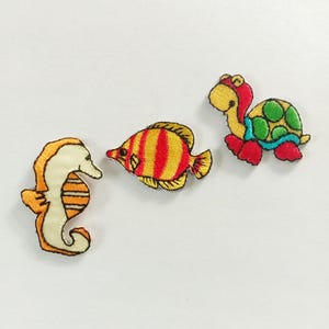 May include: Three embroidered fabric patches featuring marine life. One patch is an orange and cream seahorse, one is a yellow and red striped tropical fish, and one is a green, yellow, and red turtle with a patterned shell design.