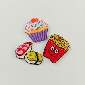 Op de afbeelding: Drie geborduurde patches met voedselontwerpen: een cupcake met wit glazuur en kleurrijke hagelslag in een paars vormpje, twee stukjes sushi met roze en gele toppings, en een rood bakje met patat met een lachend gezichtje.