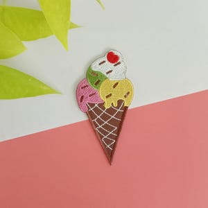 May include: Embroidered ice cream cone patch featuring three scoops: pink, green, and yellow, topped with a red cherry. The cone is brown with a waffle pattern. The patch is set against a white and pink background.