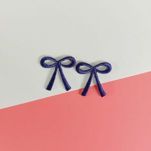 Op de afbeelding: Twee geborduurde paarse strikapplicaties. De strikken zijn symmetrisch met een lusvormige bovenkant en twee naar beneden gerichte uiteinden. De applicaties zijn geplaatst tegen een witte en roze achtergrond.