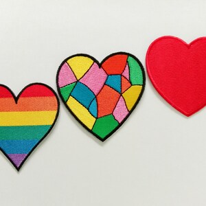Può includere: Tre toppe ricamate a forma di cuore. La prima toppa presenta strisce orizzontali rosse, arancioni, gialle, verdi, blu e viola. La seconda toppa ha un motivo multicolore a vetrata. La terza toppa è di colore rosso tinta unita.