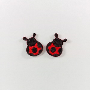 Pode incluir: Dois pequenos emblemas bordados em forma de joaninhas. Cada emblema apresenta uma cabeça e corpo pretos com um padrão de estrela vermelho vibrante nas costas, acentuado por um ponto preto central. O bordado tem um acabamento texturizado.