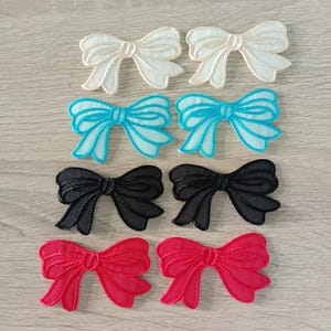 Könnte beinhalten: Acht gestickte Stoffschleifen in verschiedenen Farben. Zwei weiße Schleifen, zwei türkisfarbene Schleifen, zwei schwarze Schleifen und zwei rote Schleifen.