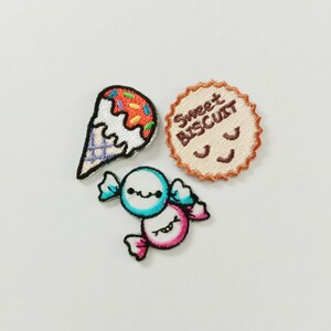 Puede incluir: Tres parches bordados que muestran un cono de helado colorido con chispas, una galleta redonda de color beige con el texto Sweet Biscuit y una cara sonriente, y un caramelo envuelto con un diseño azul y rosa y una pequeña cara sonriente.