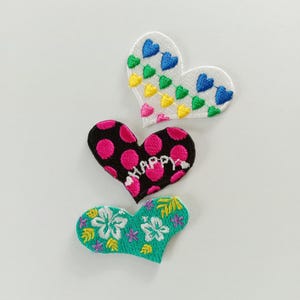 Può includere: Tre toppe ricamate a forma di cuore. La toppa in alto è bianca con una ghirlanda di piccoli cuori colorati. La toppa centrale è nera a pois rosa con la parola HAPPY ricamata in bianco. La toppa in basso è verde acqua con fiori bianchi.