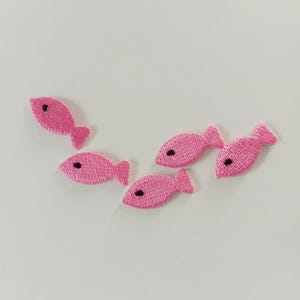 May include: Five pink embroidered fish patches with black eye details are arranged on a white background. The fish have a simple, cartoon-like design, and are likely intended for decorative use on clothing or crafts.