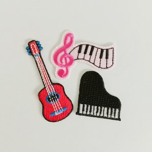 Könnte beinhalten: Drei aufgestickte Aufnäher mit Musikinstrumenten. Eine rote Gitarre mit blauen Akzenten, ein rosa Violinschlüssel mit einer Klaviertastatur und ein schwarzes Klavier mit weißen Tasten. Auf weißem Hintergrund.