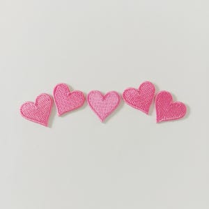 May include: Five pink heart-shaped embroidered patches arranged in a row. Each heart has a textured appearance, with a lighter pink color in the center and a slightly darker pink outline. The patches are set against a plain white background.