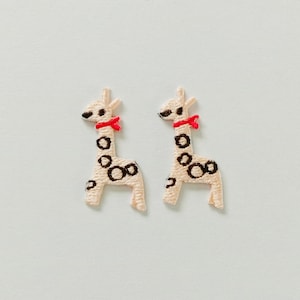 May include: Two embroidered giraffe patches. Each giraffe has a cream-colored body with black spots, a red bow, and black eyes. The patches are likely for clothing or craft projects.