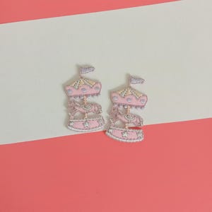 Op de afbeelding: Twee geborduurde carrouselpatches. Elke patch heeft een roze carrousel met een gouden dak, een paard en een kleine vlag. De patches zijn geplaatst tegen een roze en witte achtergrond.