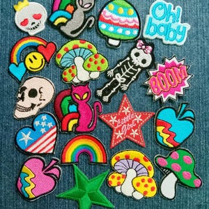 May include: A collection of embroidered patches in various shapes and colors. Designs include a skull with a crown, cats, mushrooms, rainbows, hearts, stars, and the text "Oh! baby" and "BOOM". The patches are on a blue denim background.