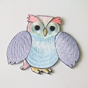 May include: Embroidered owl patch in pastel colors. The owl features a light blue body, lavender wings, and a pink and peach head. The owl's eyes are black with gold accents, and it has yellow feet. This patch is suitable for clothing or accessories.