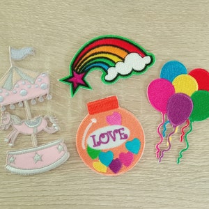 May include: Five colorful iron-on patches. A pink carousel with a white horse, a rainbow with a star, a jar with the word "LOVE" and hearts, and a cluster of balloons in red, blue, purple, yellow, and green.