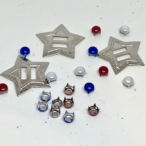 May include: An assortment of decorative craft supplies, including silver star-shaped embellishments and small, colorful, round-topped studs in red, white, and blue. These items are scattered on a white surface.