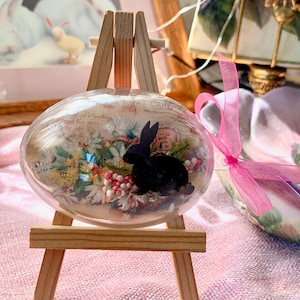 Könnte beinhalten: Ein klarer, eiförmiger Glasornament mit einer schwarzen Kaninchensilhouette im Inneren, präsentiert auf einer kleinen Holzstaffelei. Das Ornament enthält florale Elemente und ist mit einem rosa Band gebunden. Im Hintergrund ist ein Gemälde von Kaninchen zu sehen.