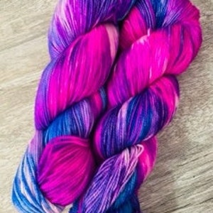 Könnte beinhalten: Zwei Knäuel Garn in einer leuchtenden pink-, lila- und blauen Farbkombination mit einem weißen Wirbelmuster.