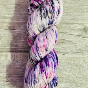 Könnte beinhalten: Eine handgefärbte Garnrolle in weißer Basis mit Spritzern in Pink, Lila und Schwarz.